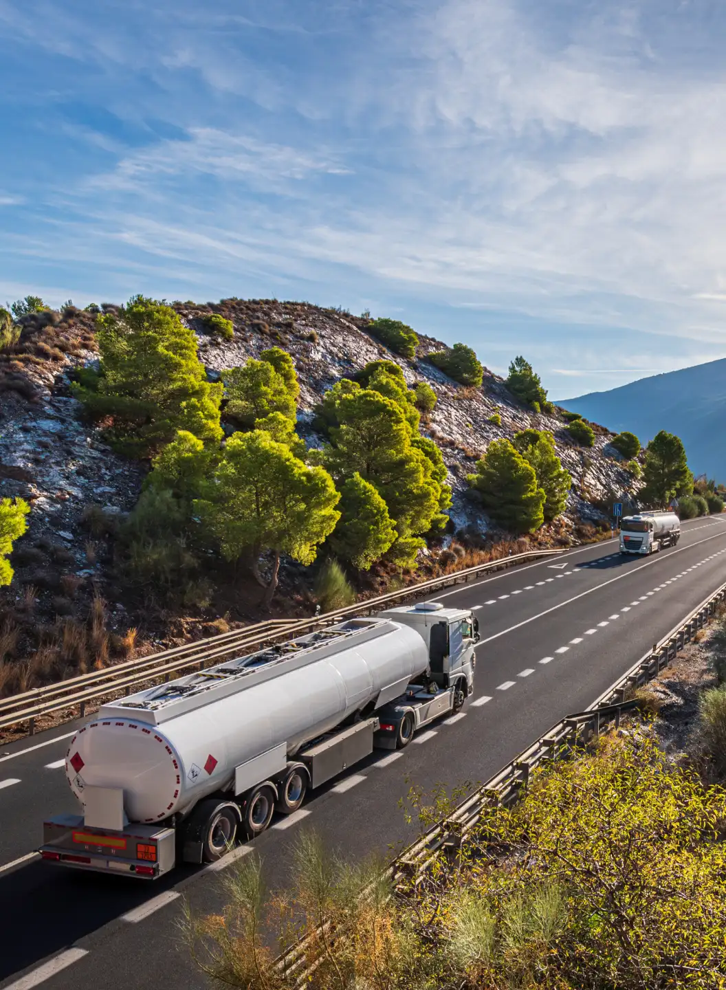Two silver tanker trucks driving on a highway next to a rocky hillside with green trees.