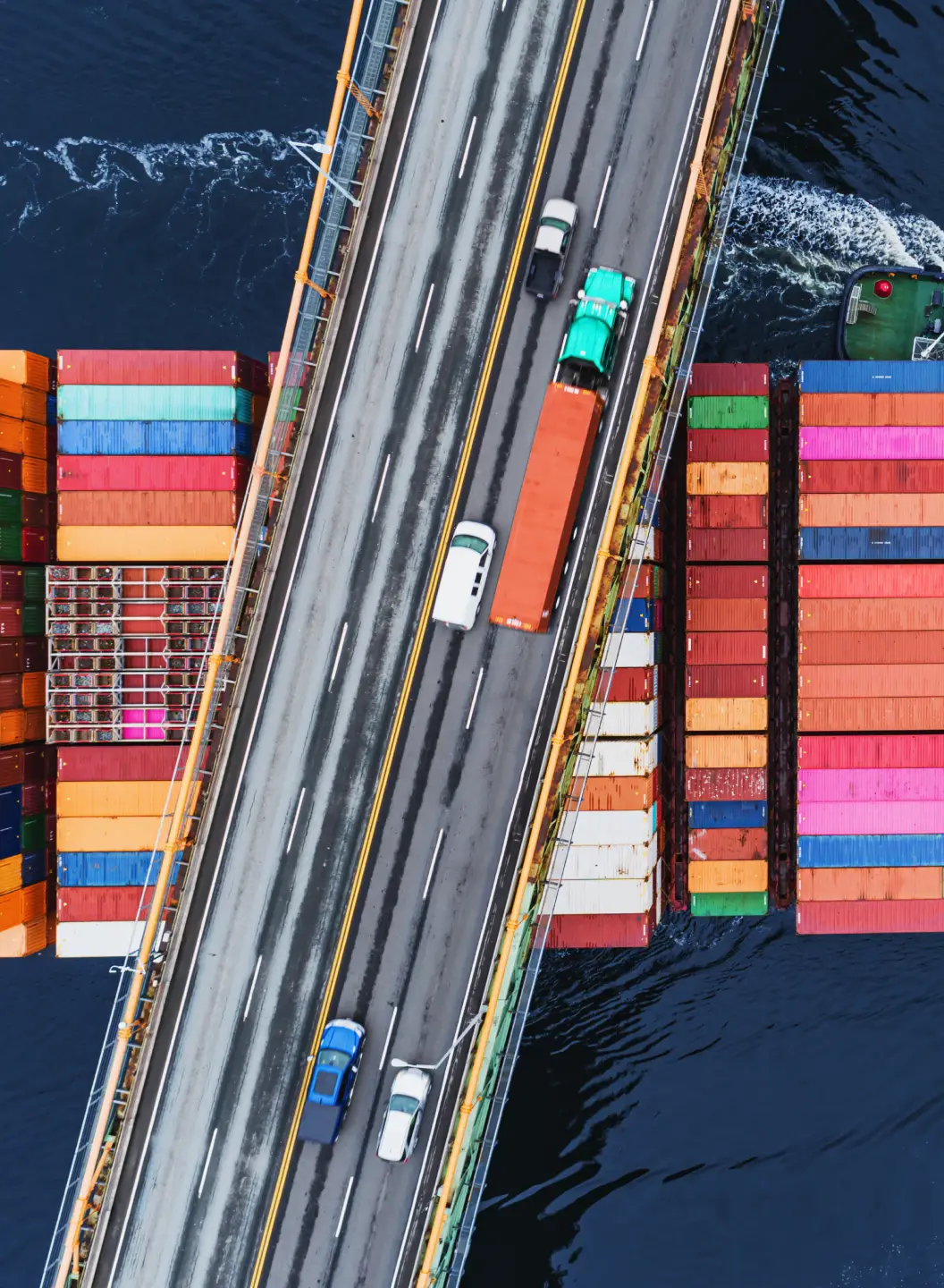 Overhead view of a bridge with cars and a truck crossing over water beside stacked shipping containers.