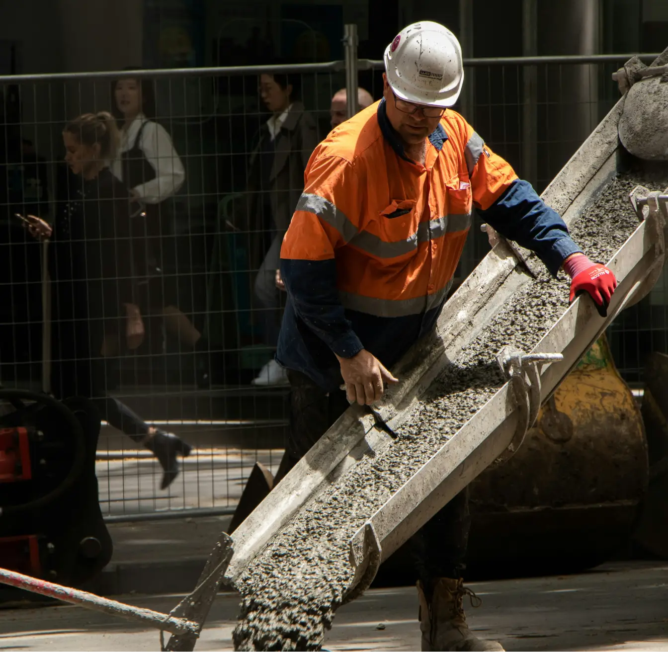 Construction worker in orange safety gear guiding wet concrete from a chute on a worksite.