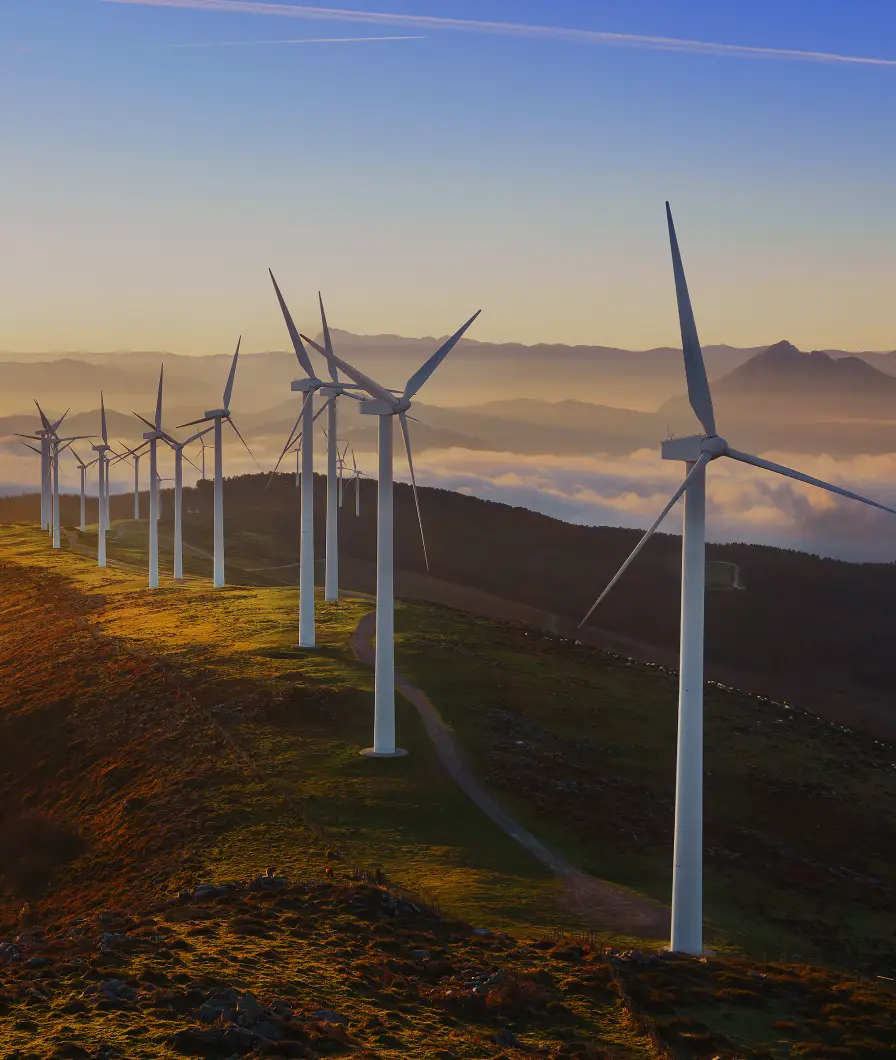 Row of wind turbines on a ridge at sunset with mist and mountains in the background.