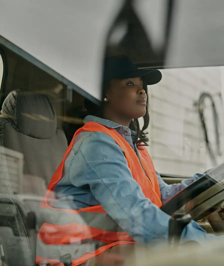 Woman wearing a black cap and orange safety vest driving a vehicle, seen through the window.