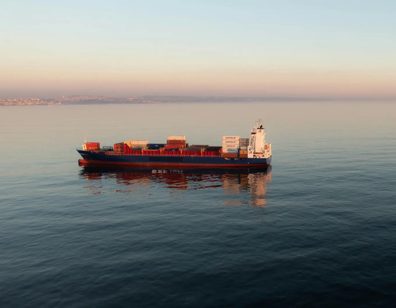 Cargo ship with containers sailing on calm water at sunrise with city skyline in the background.
