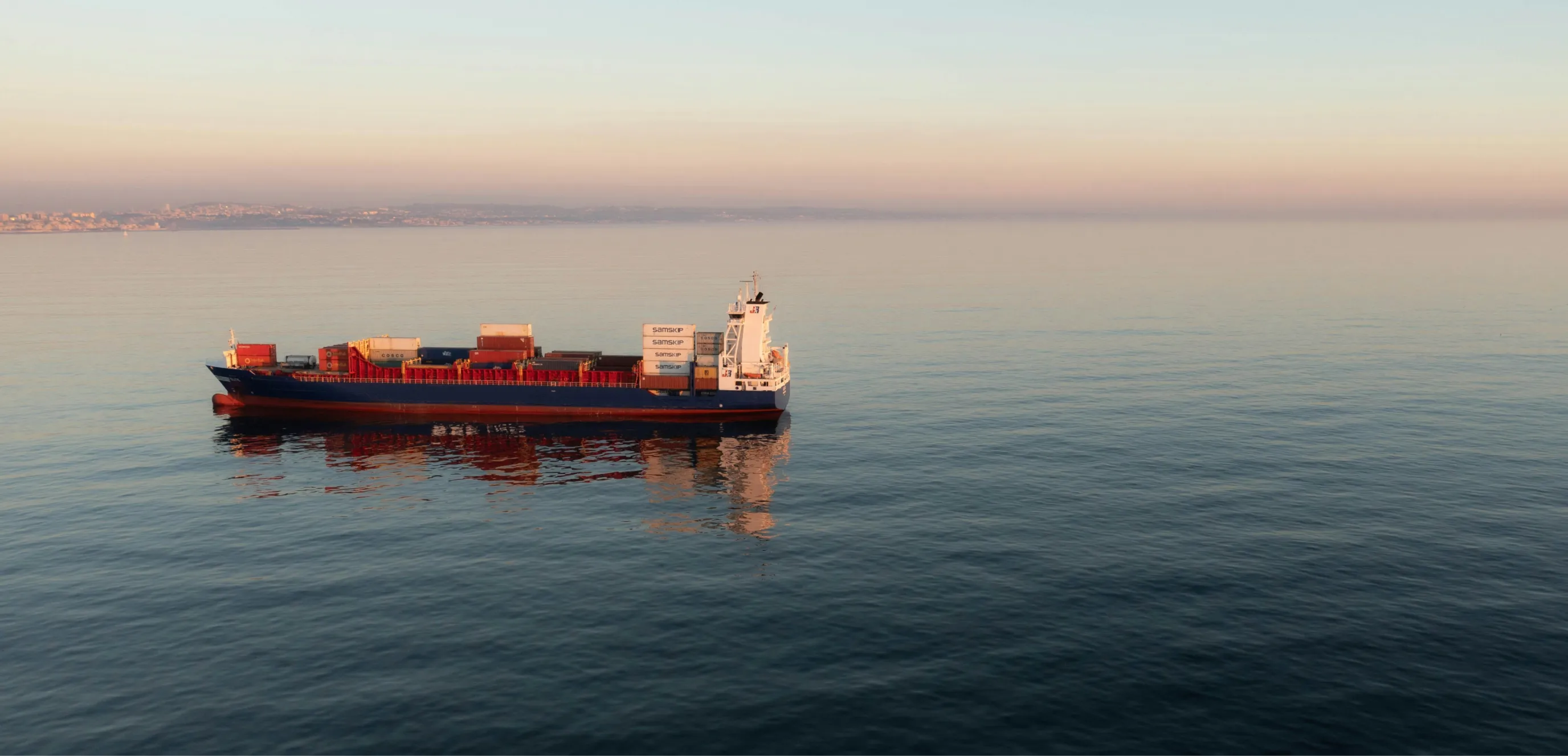 Cargo ship with containers sailing on calm sea at sunset with coastal city in the background.
