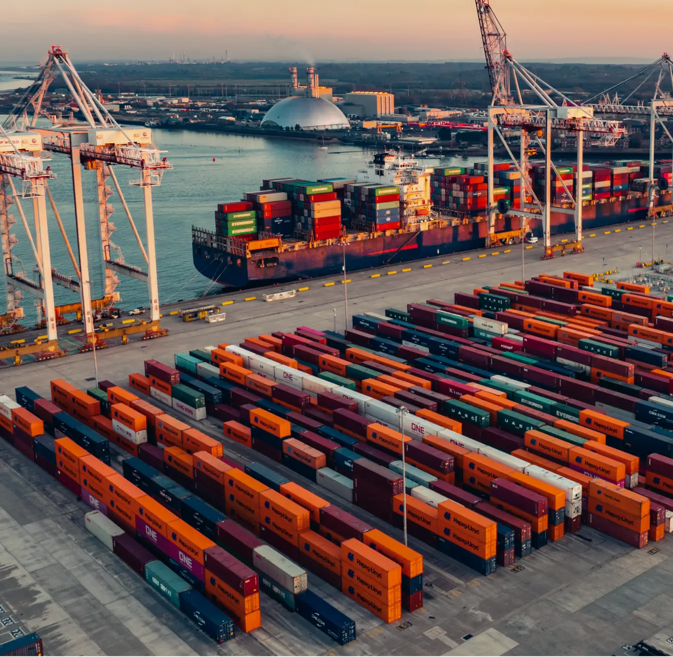 Shipping yard with stacked cargo containers and cranes unloading a large container ship in the harbor.