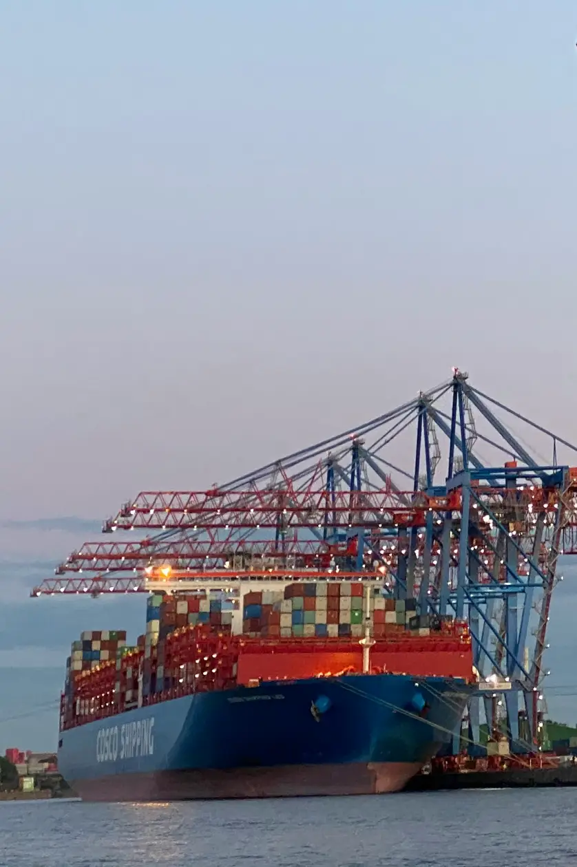 Large container ship docked at port with colorful stacked shipping containers and cranes.