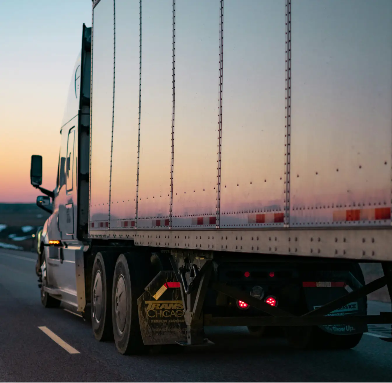 Large white semi-truck driving on highway at sunset with trailer reflecting the sky colors.
