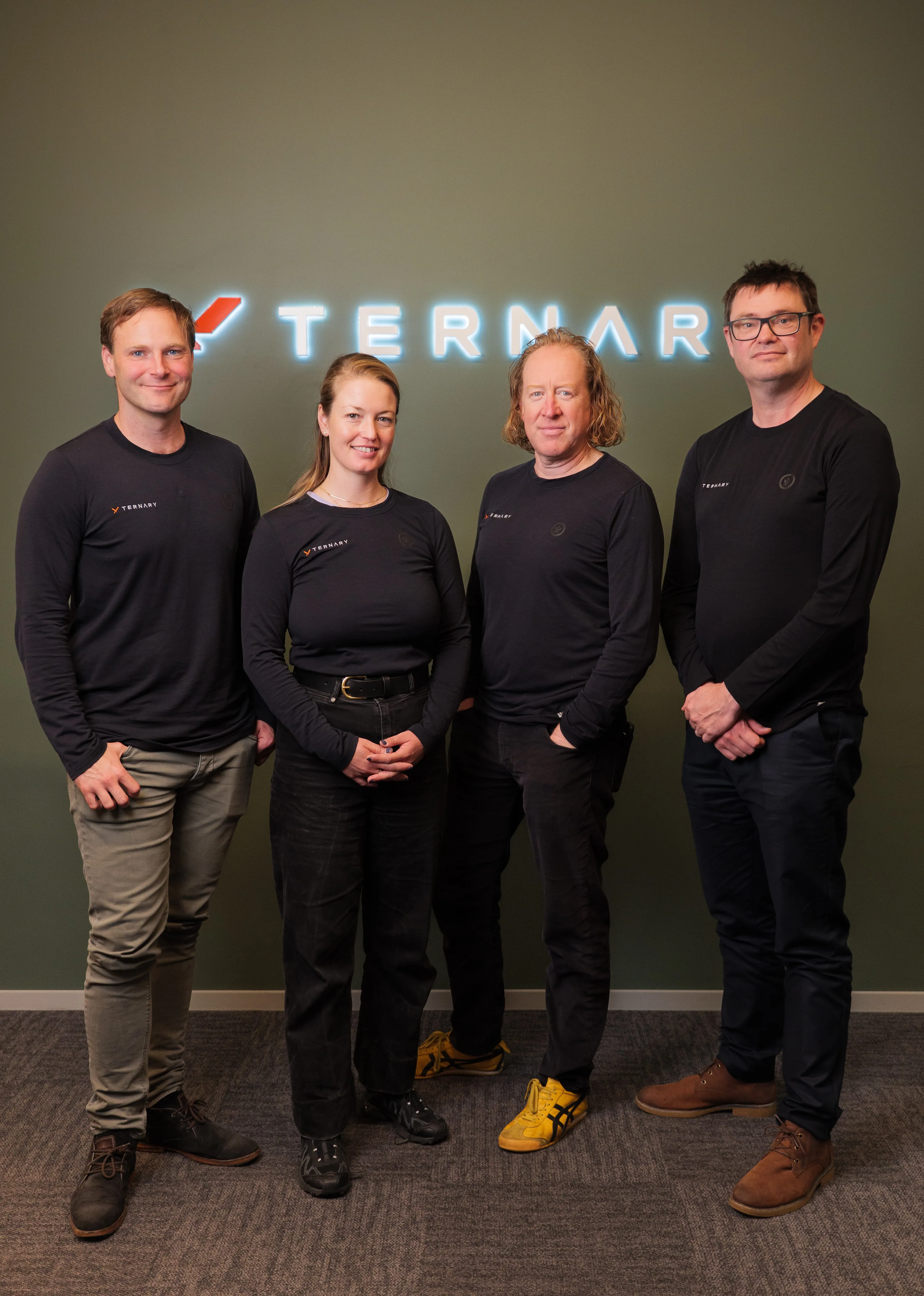 Four people wearing black Ternary shirts standing in front of a green wall with a lit Ternary sign.