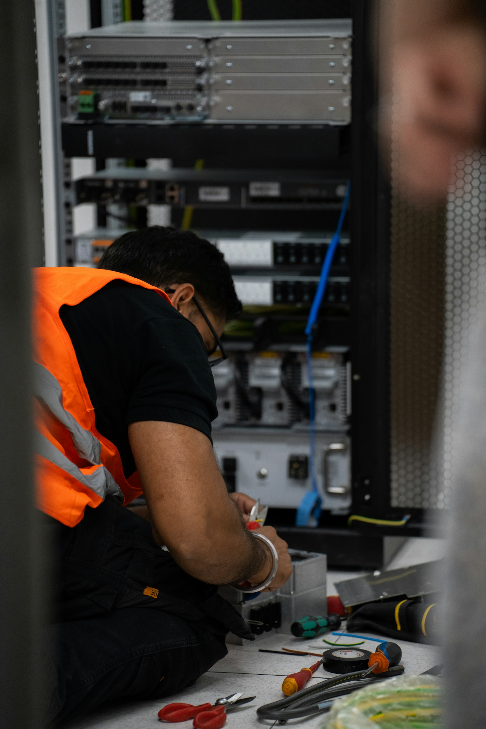 Technician in an orange hi-vis vest working on equipment beside a server rack.