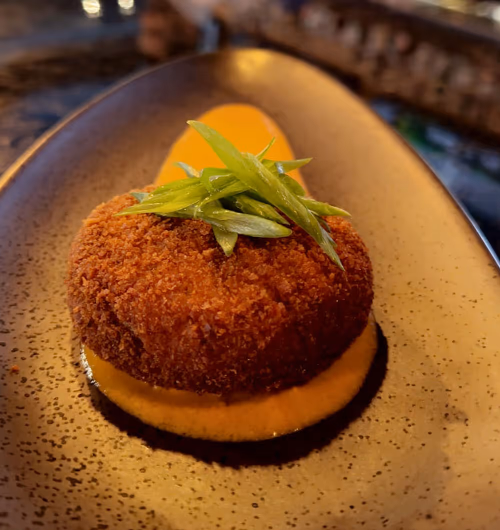Breaded round fried food on orange sauce garnished with green sliced herbs on a black plate.
