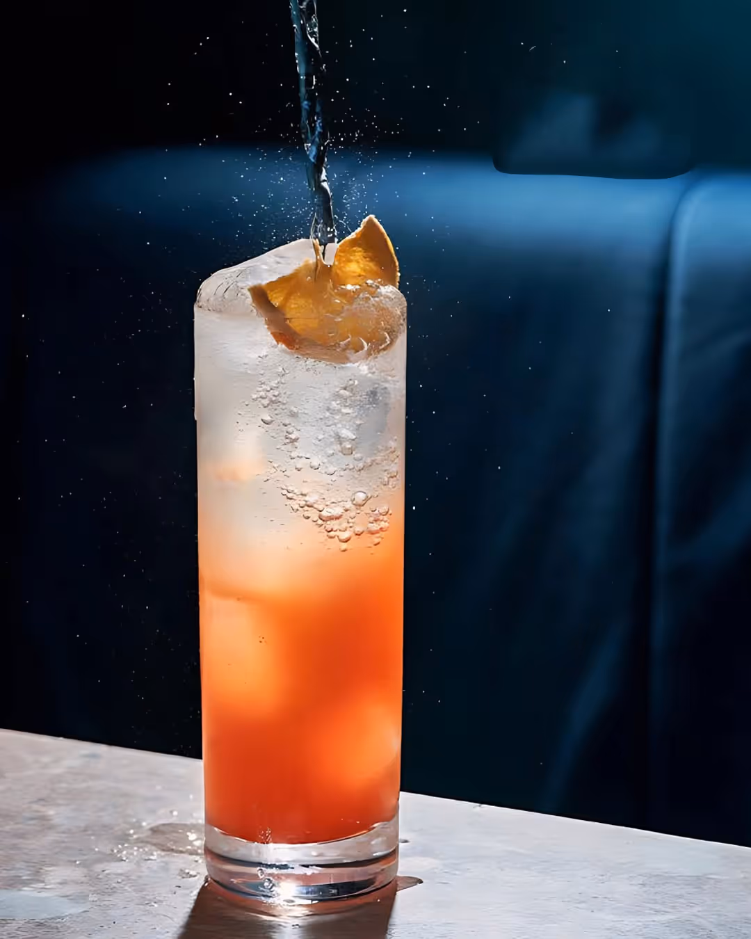 Tall glass with orange and clear layered drink, ice cubes, and orange peel garnish with liquid pouring in.