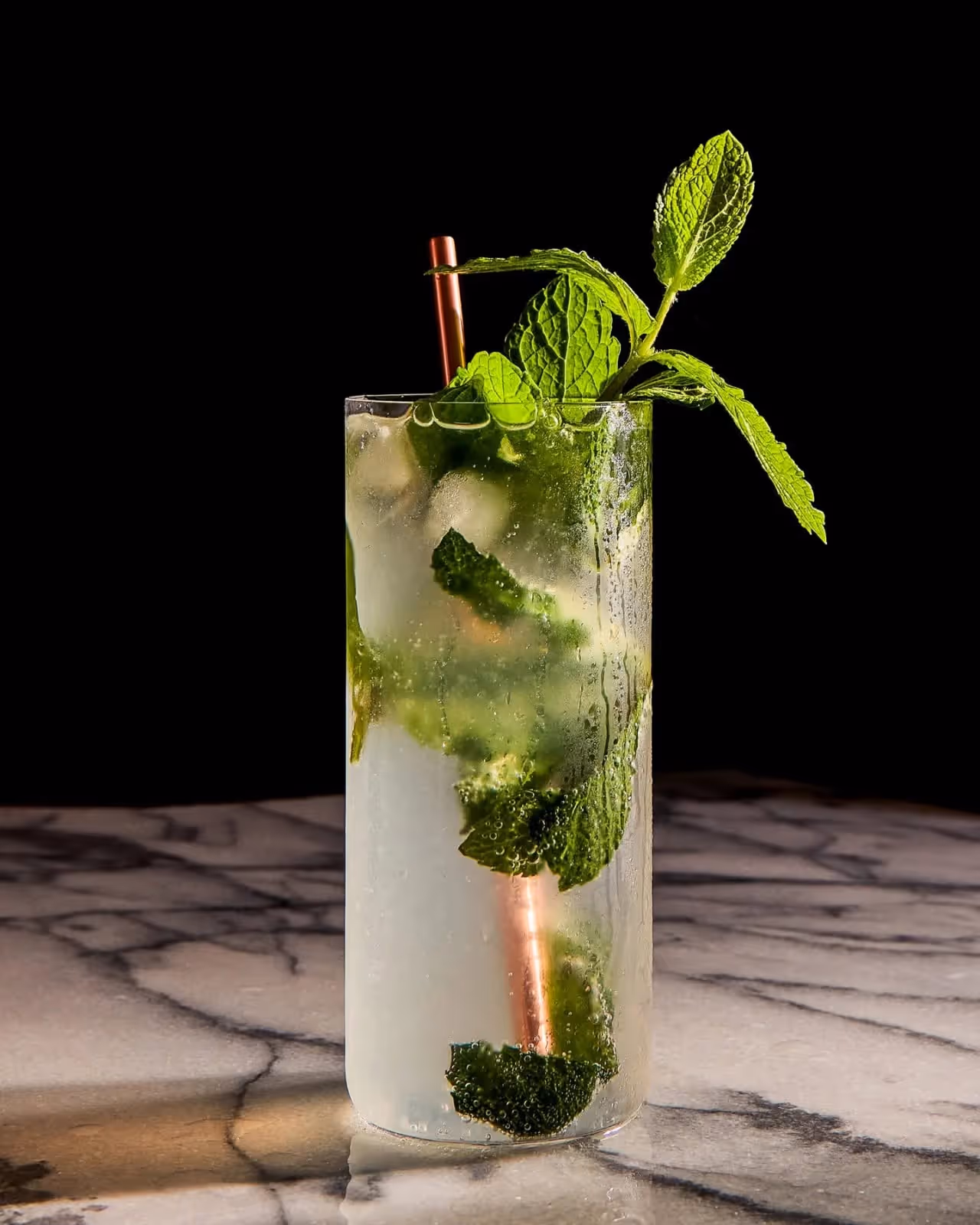 Tall glass of mojito cocktail with fresh mint leaves, ice, and a copper straw on a marble surface.