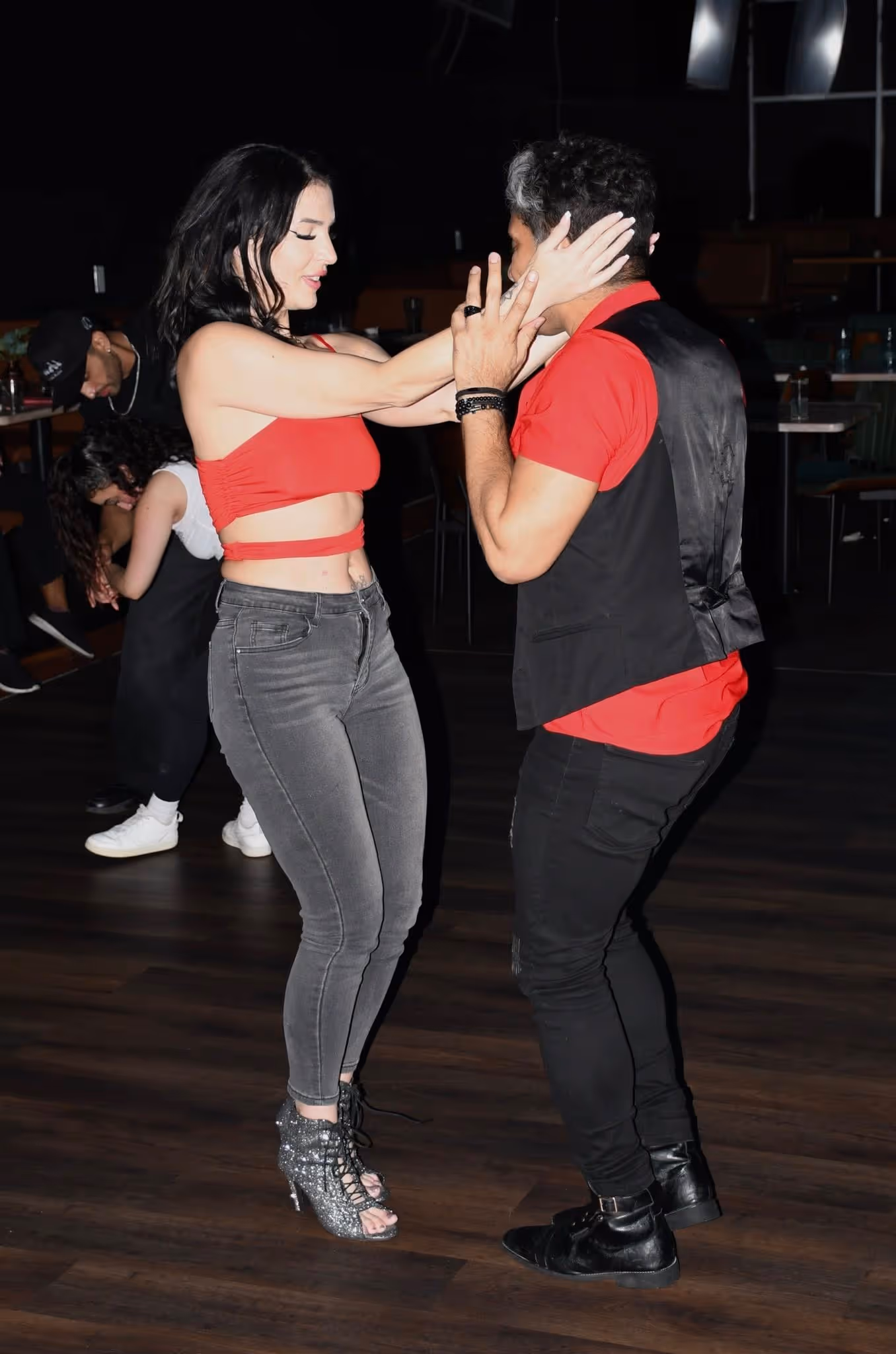 A man and woman dance closely in a dimly lit room with wooden floors and other dancers in background.
