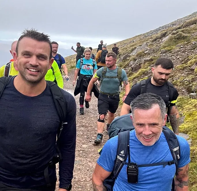 Group of hikers smiling and walking on a rocky mountain trail under cloudy sky.
