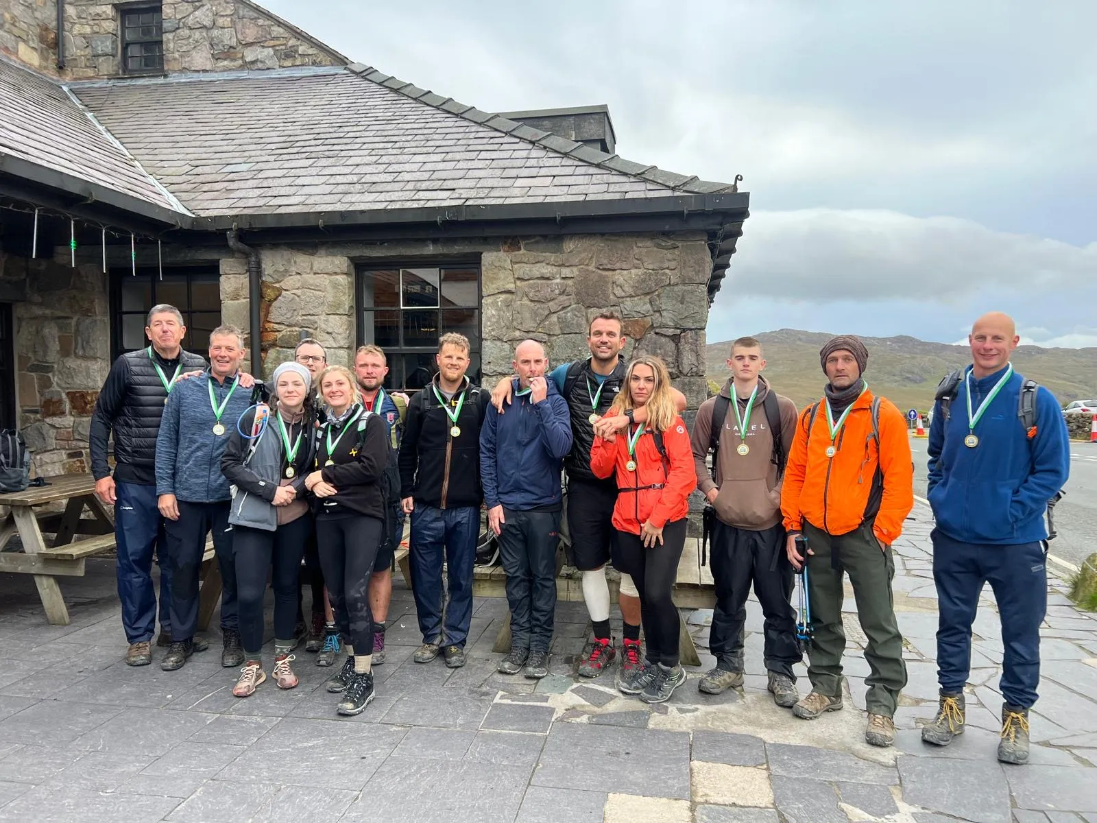 Group of hikers posing with medals outside a stone building under a cloudy sky.