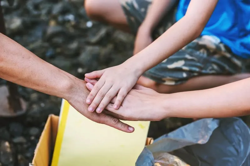 Three hands stacked in a gesture of teamwork or support over a yellow box outdoors.