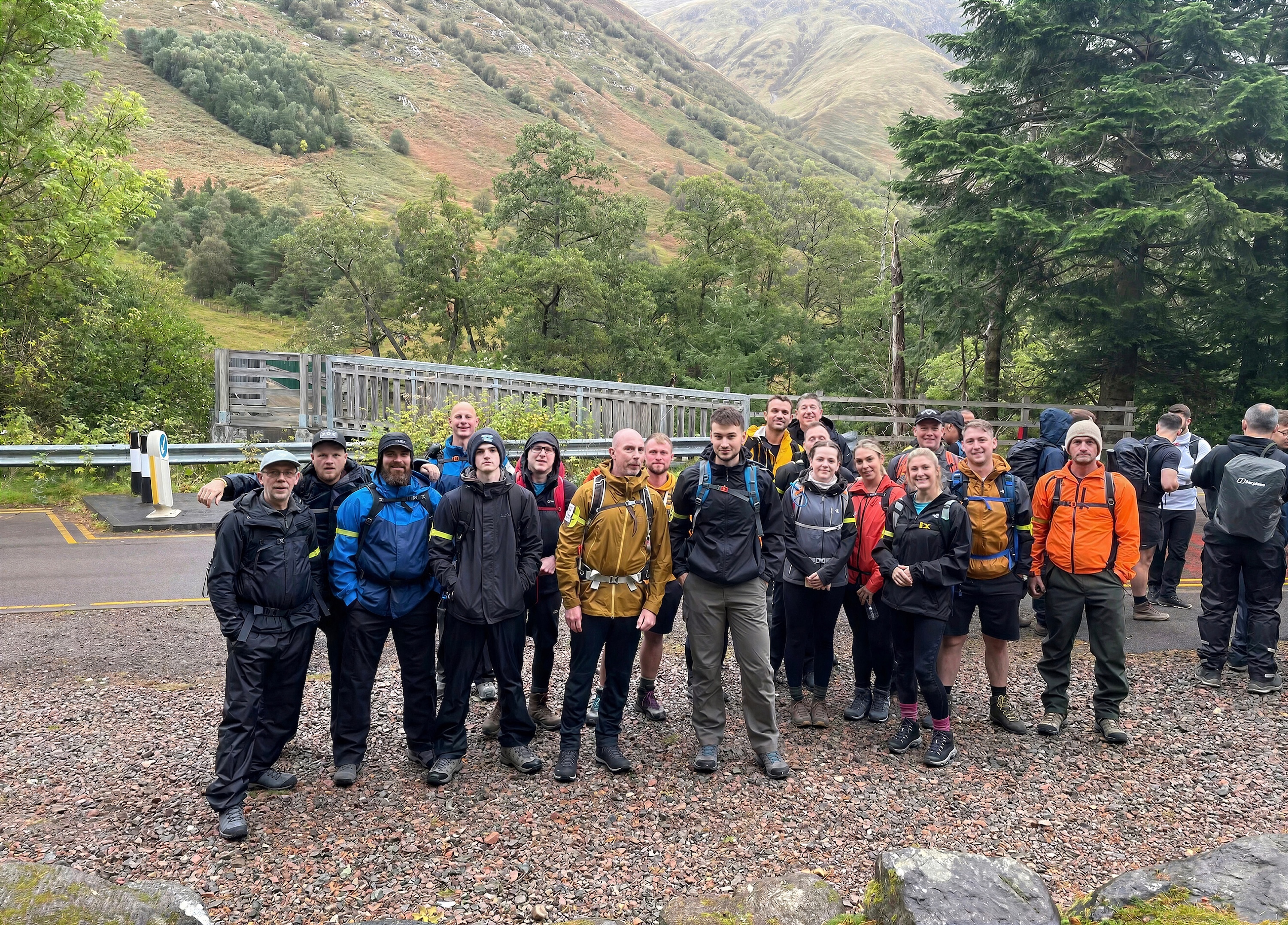 Group of people dressed in hiking gear standing on a gravel area with trees and a wooden fence behind.