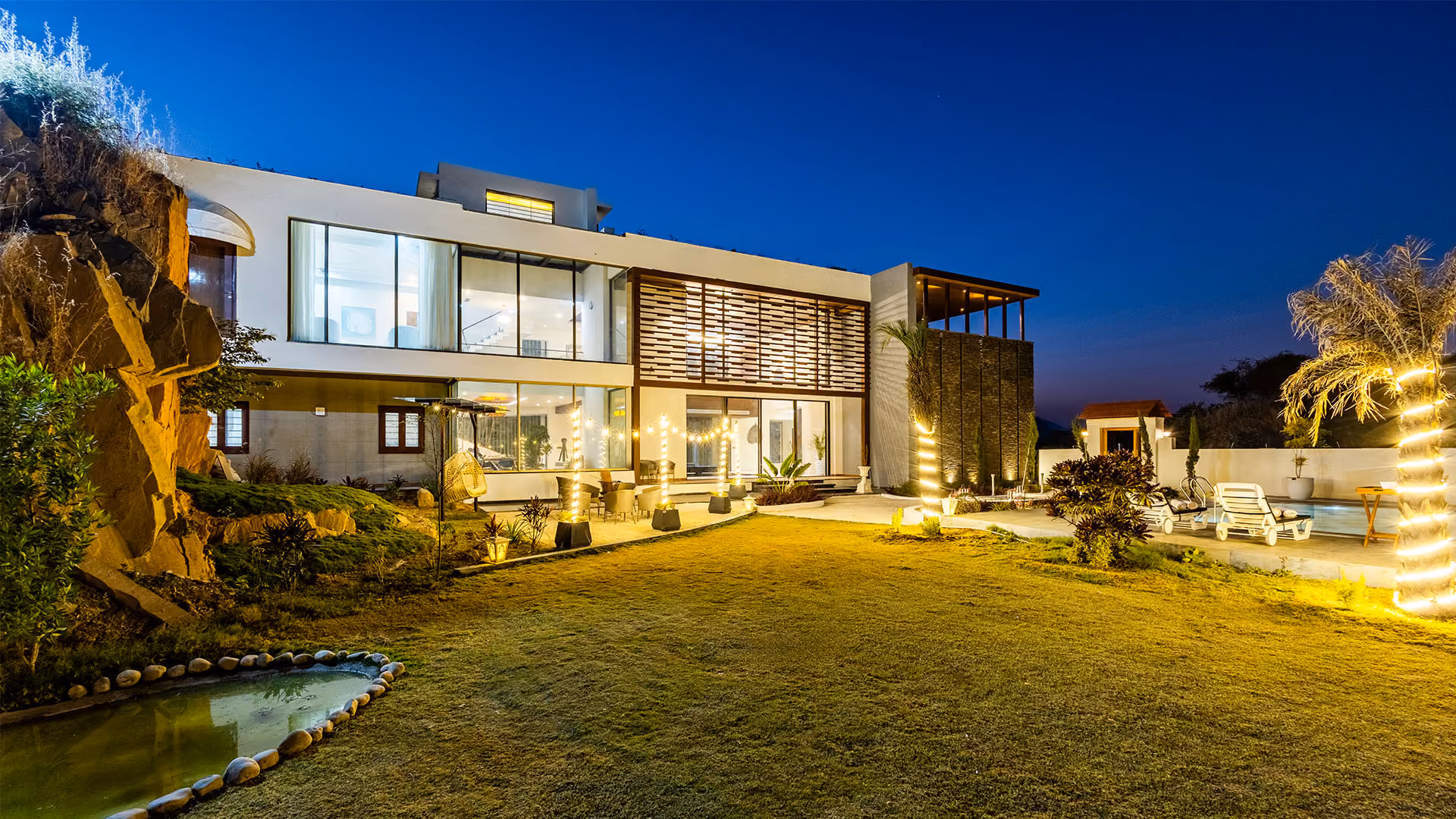 Modern mansion at dusk with lit outdoor patio, lawn, pool, and palm trees wrapped in lights.