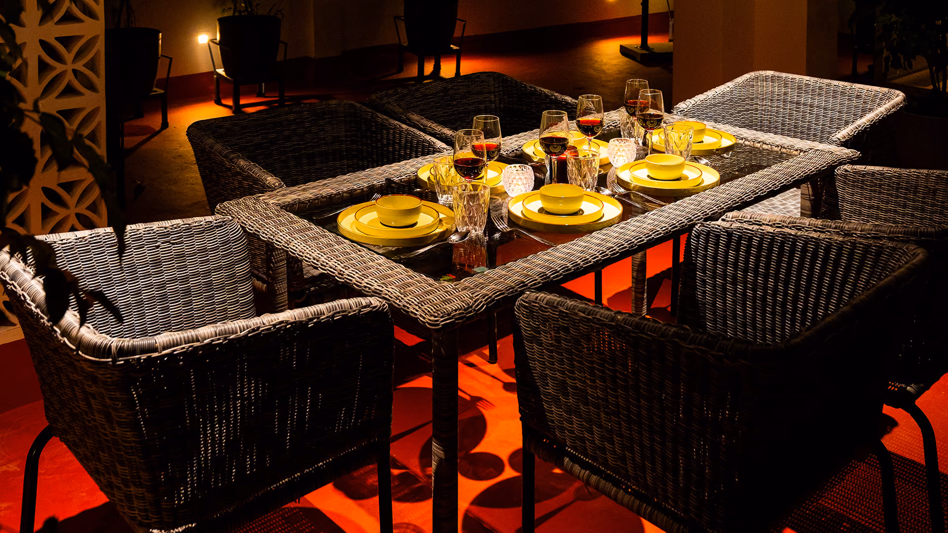 Outdoor wicker dining table set for six with yellow plates and glasses of red wine under warm lighting.