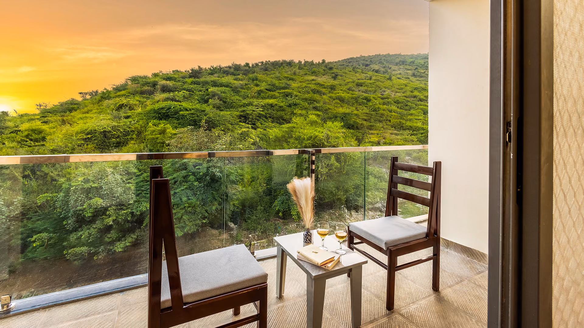 Balcony with two chairs, a table holding two glasses, books, and a vase, overlooking green hills at sunset.