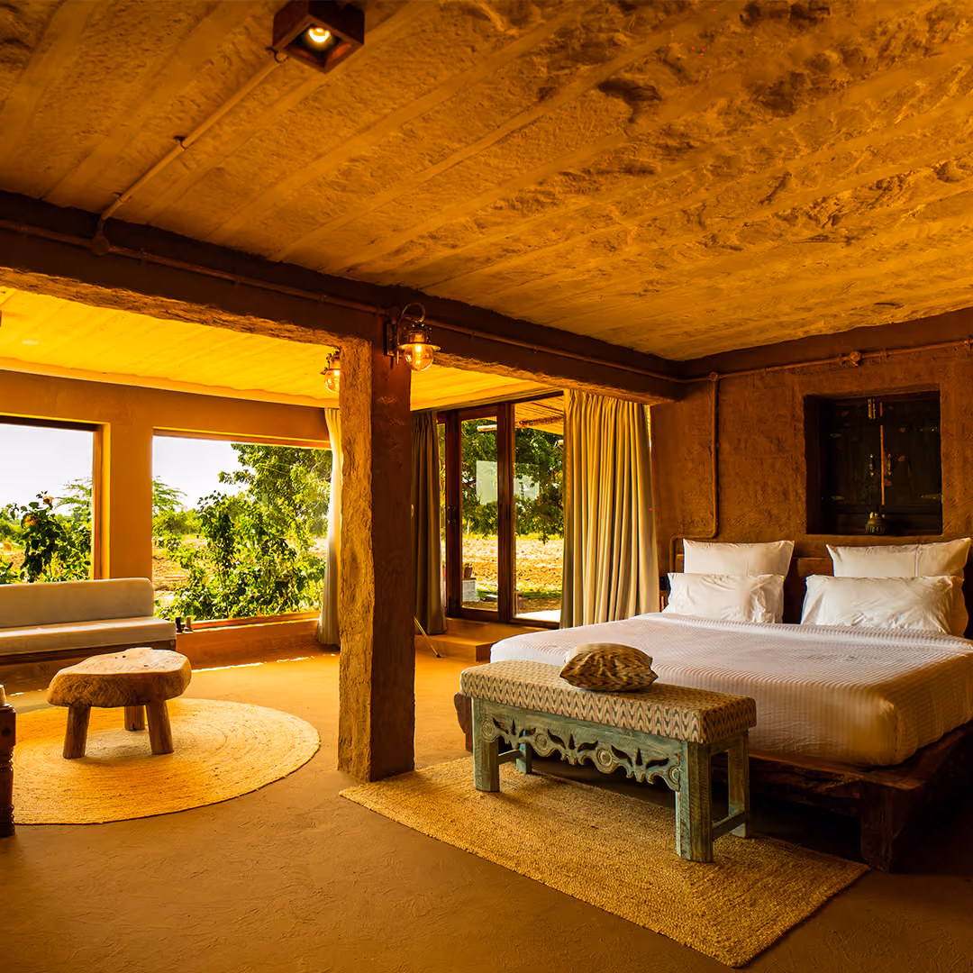 Cozy rustic bedroom with large bed, wooden bench, natural textures, and windows showing greenery outside.