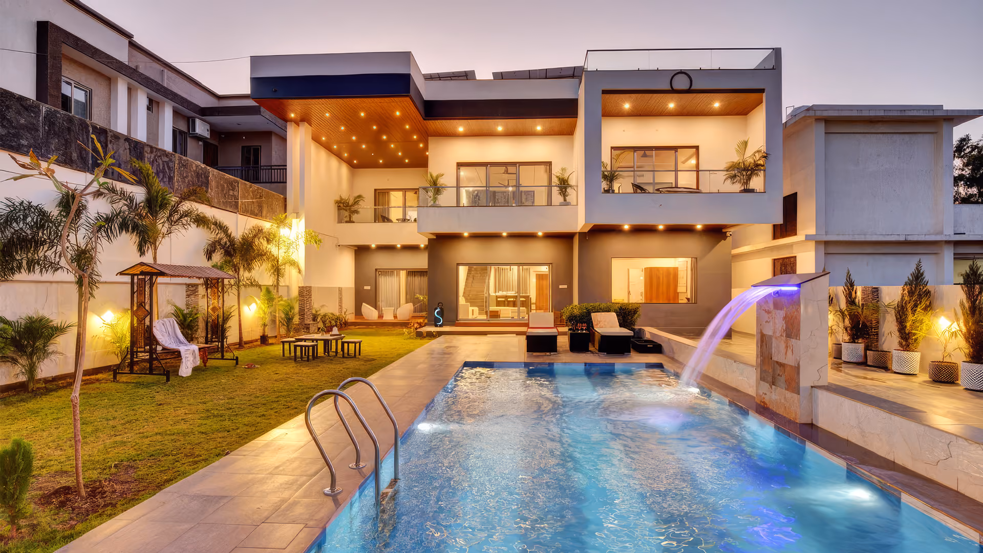 Modern two-story house with illuminated outdoor swimming pool and lawn with seating and plants.