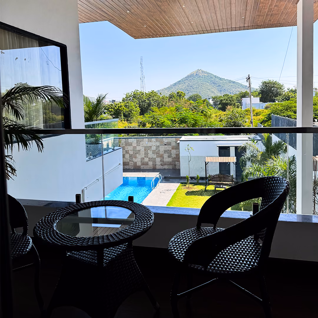 Balcony with black wicker chairs and table overlooking a pool, garden, and hill under blue sky.