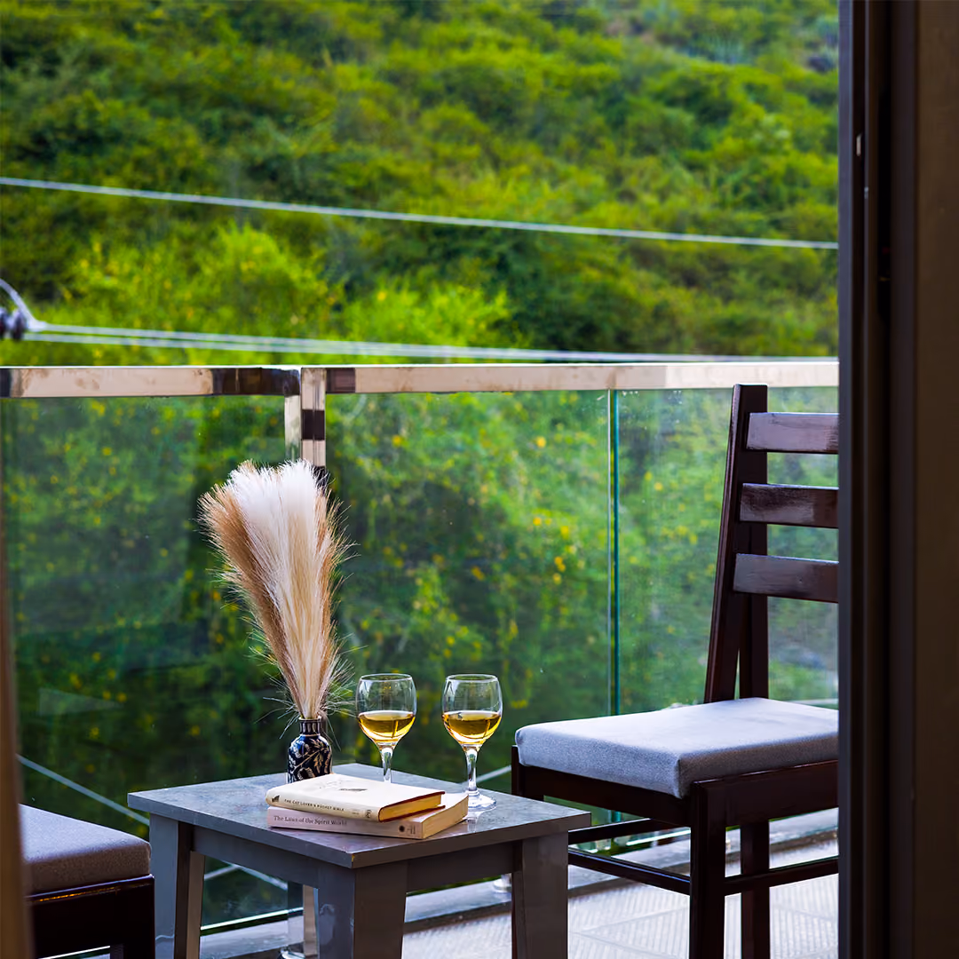 Balcony table with two glasses, books, and a vase with dried grass, overlooking green hills.
