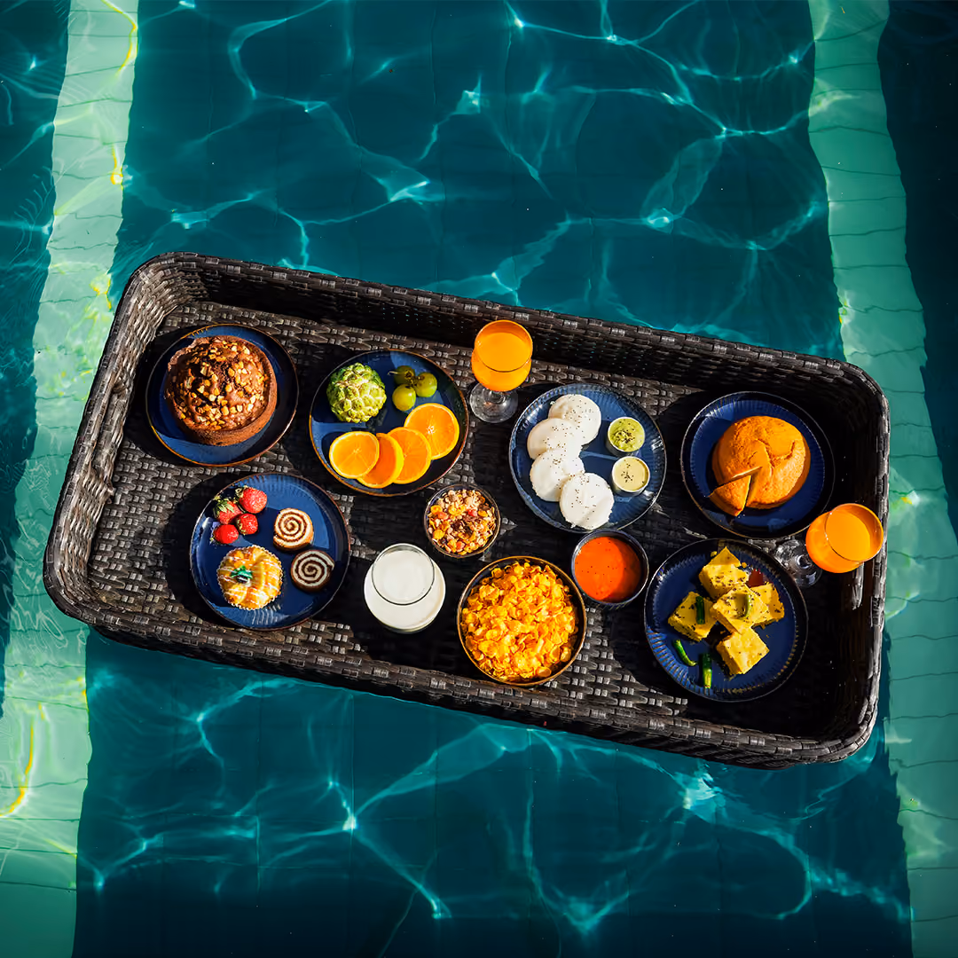 Floating basket in pool holding plates of fruit, snacks, two glasses of juice, and a glass of milk.