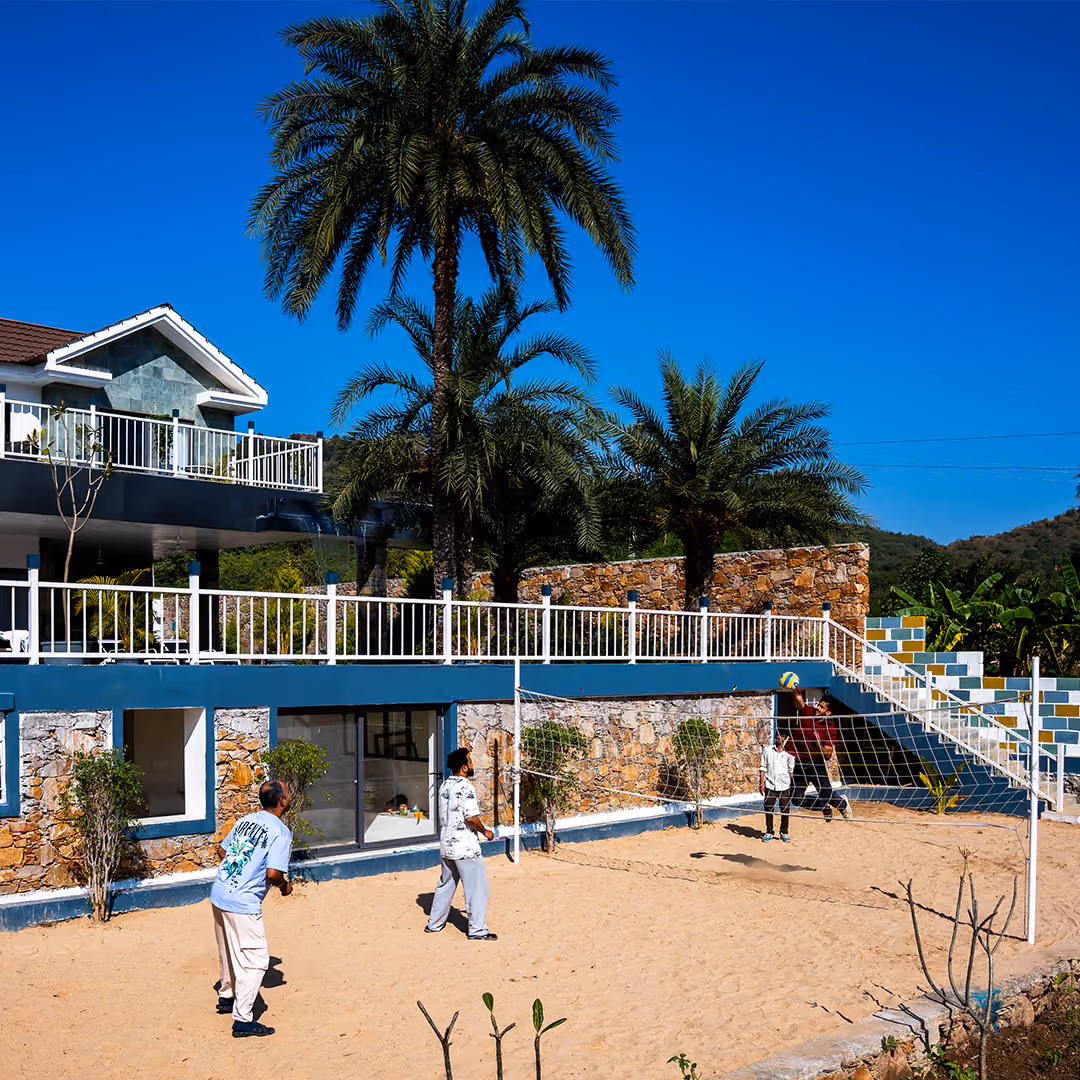 Four men playing volleyball on a sandy court beside a stone building with palm trees and clear blue sky.