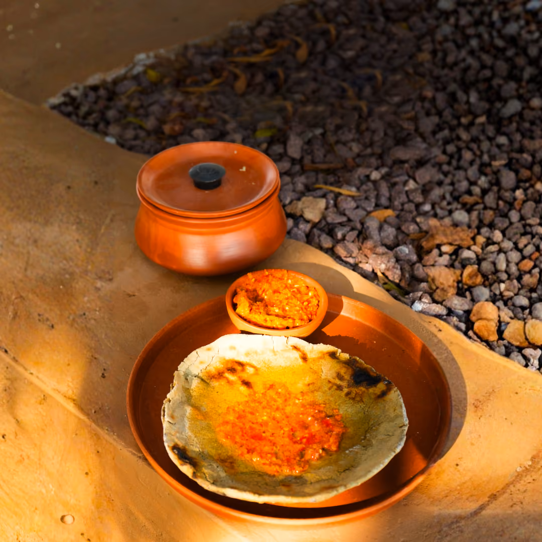 Clay plate with flatbread topped with red chutney, small bowl of chutney, and a covered clay pot.