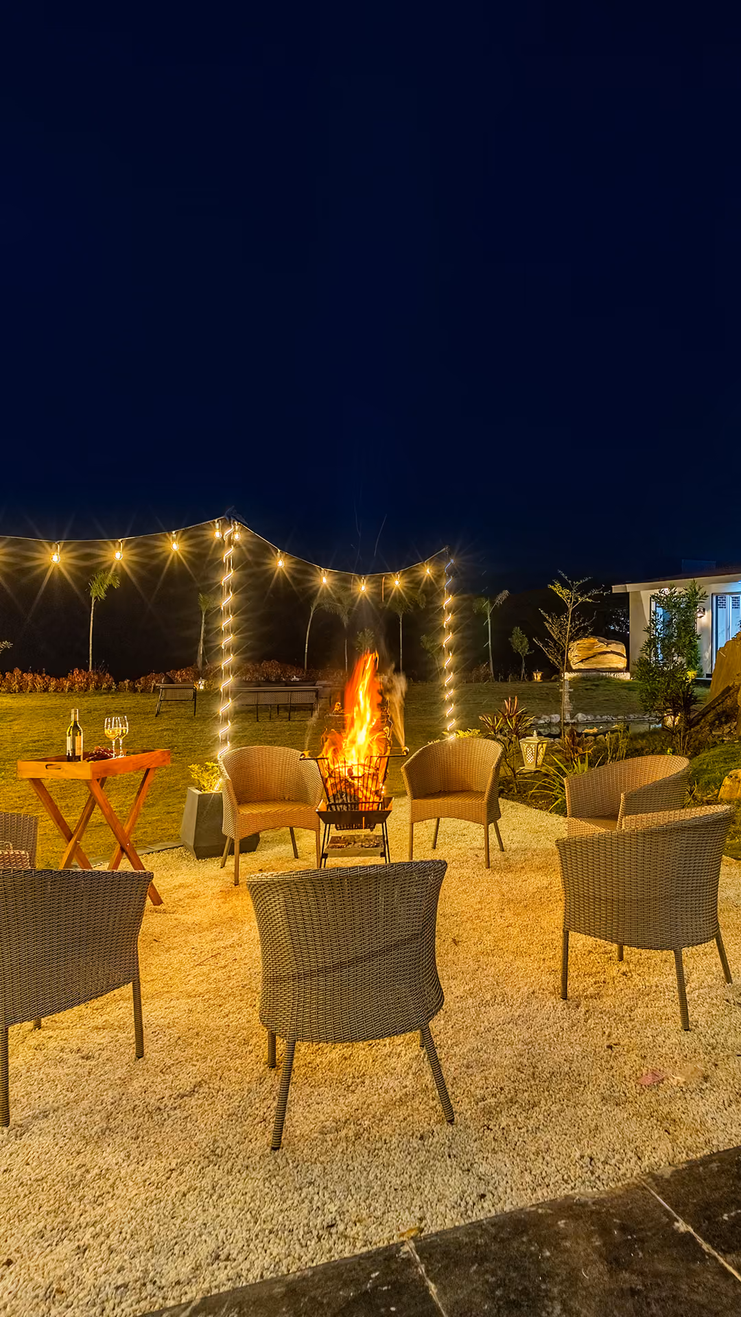 Outdoor seating area at night with wicker chairs around a lit firepit and string lights overhead.