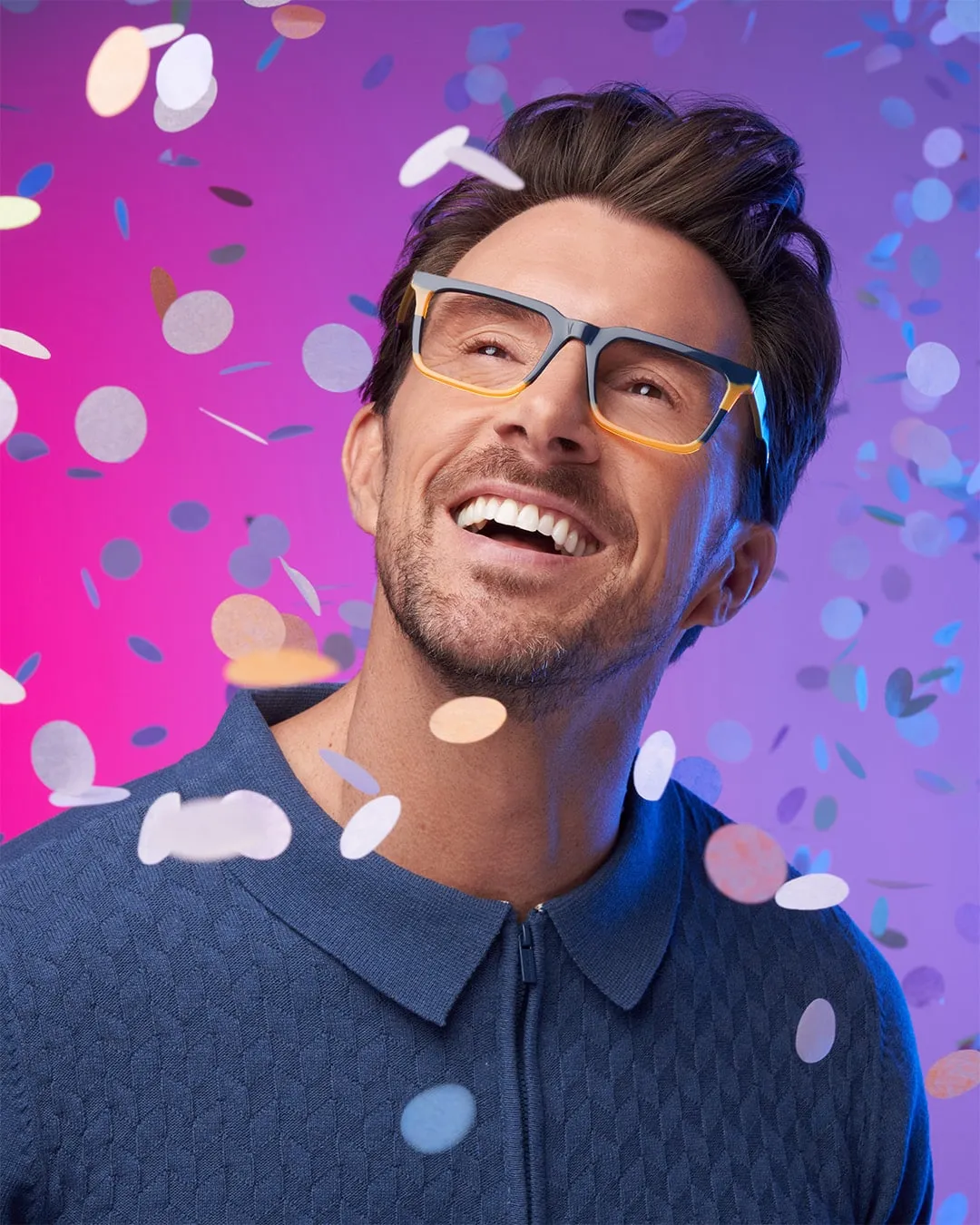Smiling man wearing glasses and a blue sweater with colorful confetti falling around him.