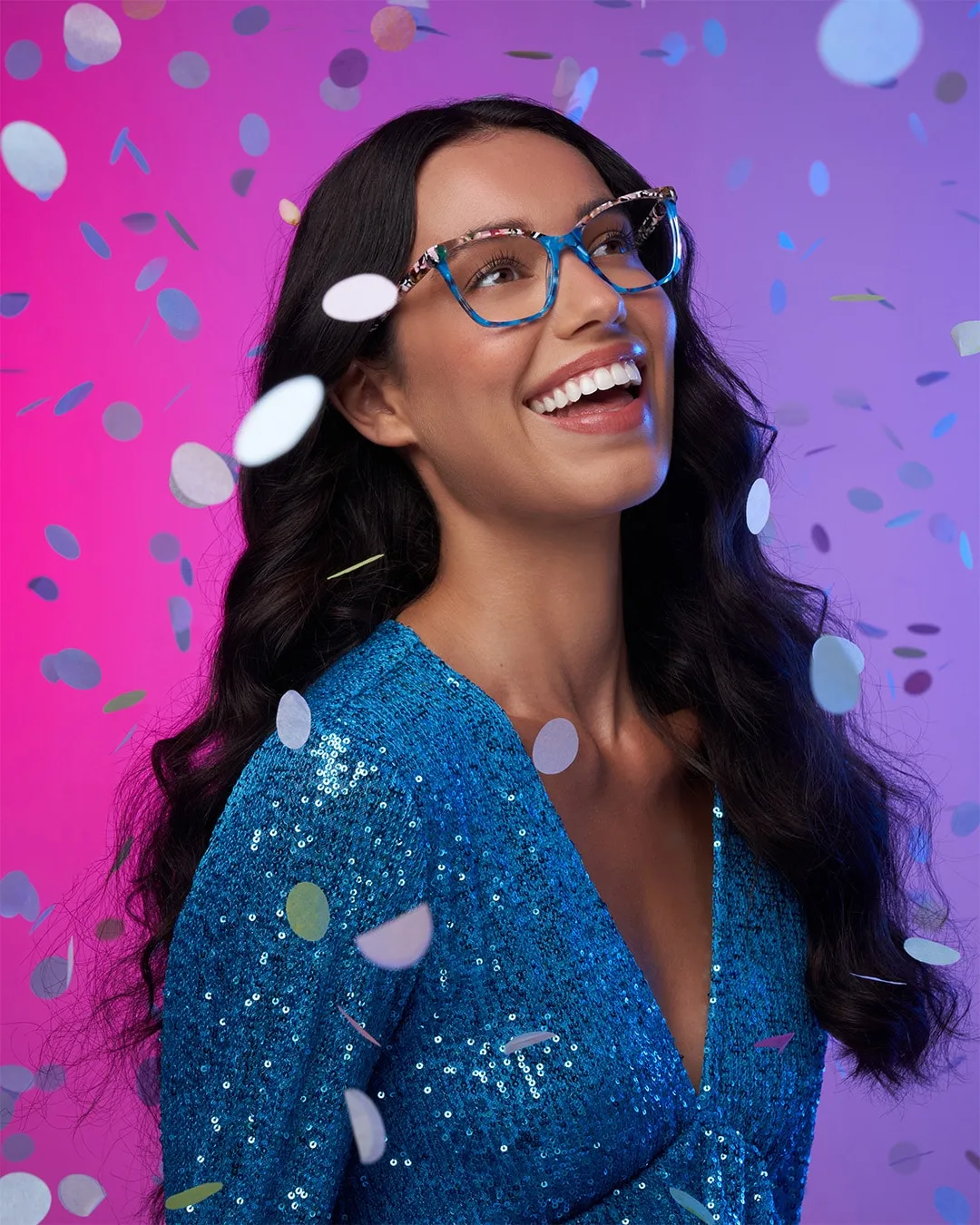 Smiling woman with long dark hair wearing blue sequin dress and glasses with confetti falling around her.
