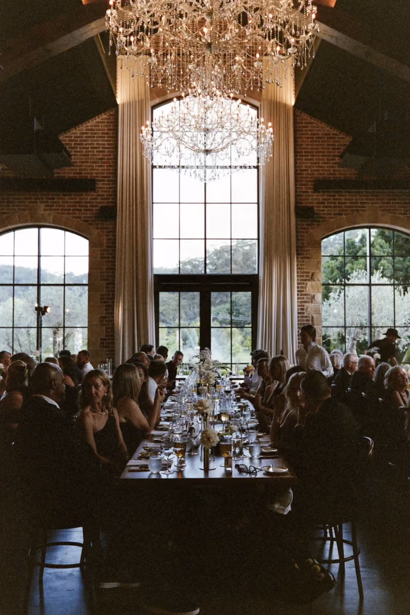 People seated at a long dining table under grand chandeliers by large windows in a brick-walled room.