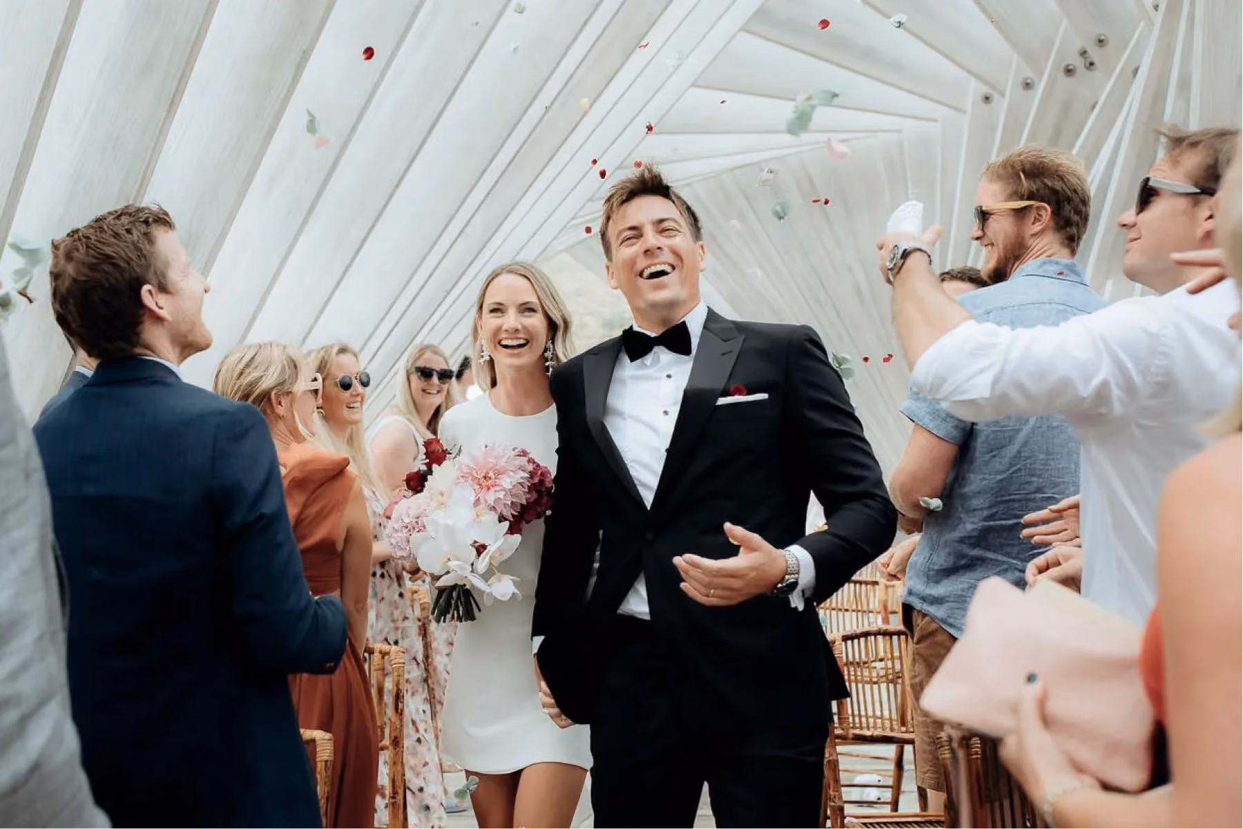 Bride in white dress and groom in tuxedo smiling and walking amid guests throwing flower petals.
