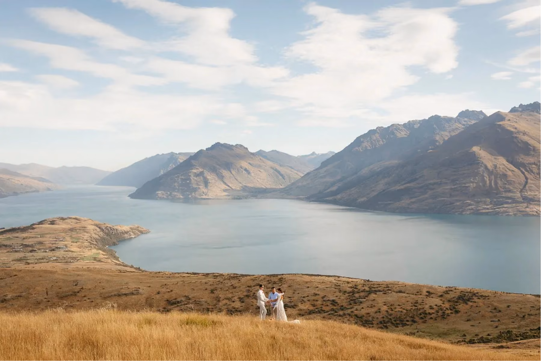 Wedding couple and officiant standing on dry grassy hill with lake and mountains in background.