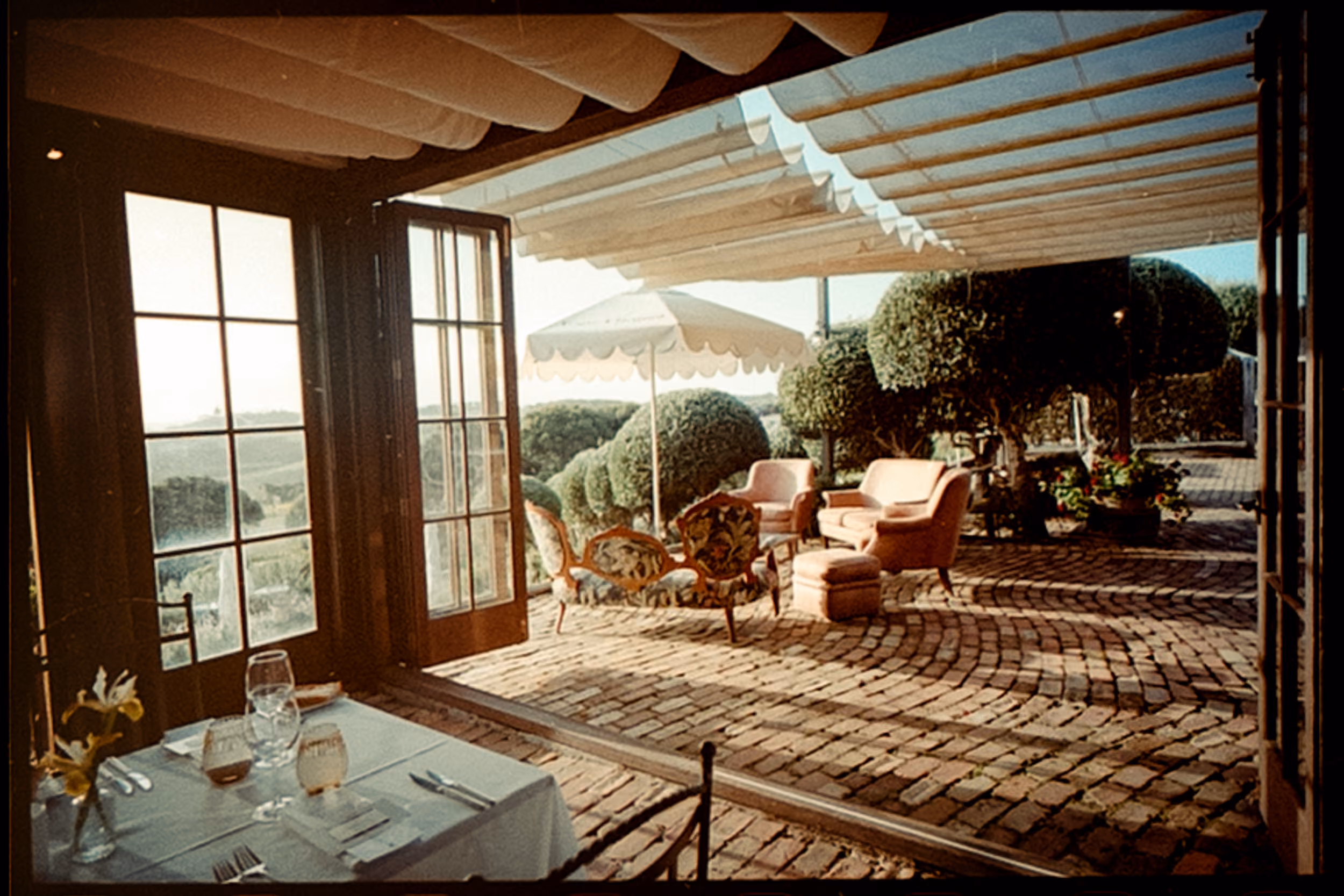 Cozy patio with cushioned chairs, umbrella, potted plants, and view through open glass doors.