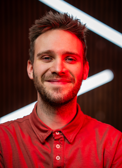 Smiling man with beard wearing black jacket with Rümpel Welt logo on green background.