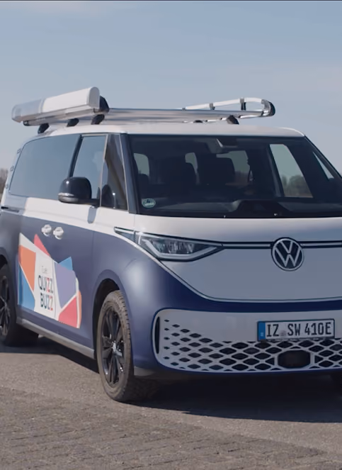 Blauer VW ID. Buzz mit Dachgepäckträger, seitliches Branding "QUIZZ BUZZ", geparkt auf Asphalt.