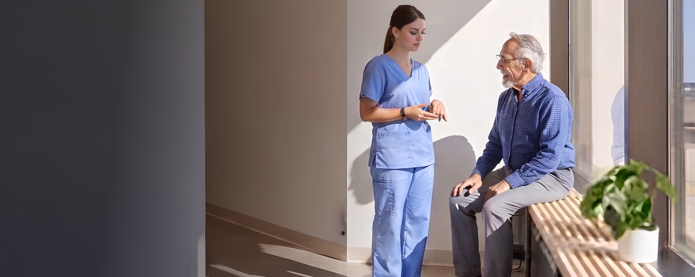 Krankenschwester spricht mit älterem Mann, der auf Fensterbank in hellem Raum sitzt.