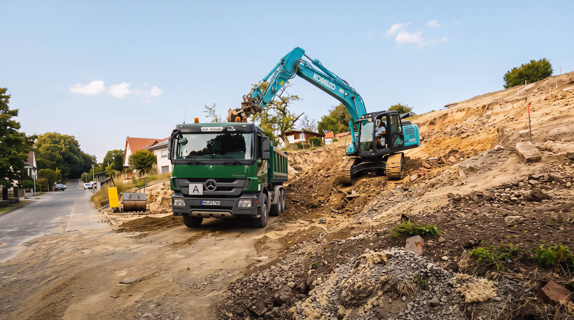 16 Tonnen Bagger lädt Erde in grünen Gebr. Schade Lastwagen auf einer Baustelle neben einer Straße mit Häusern in Borken (Hessen).