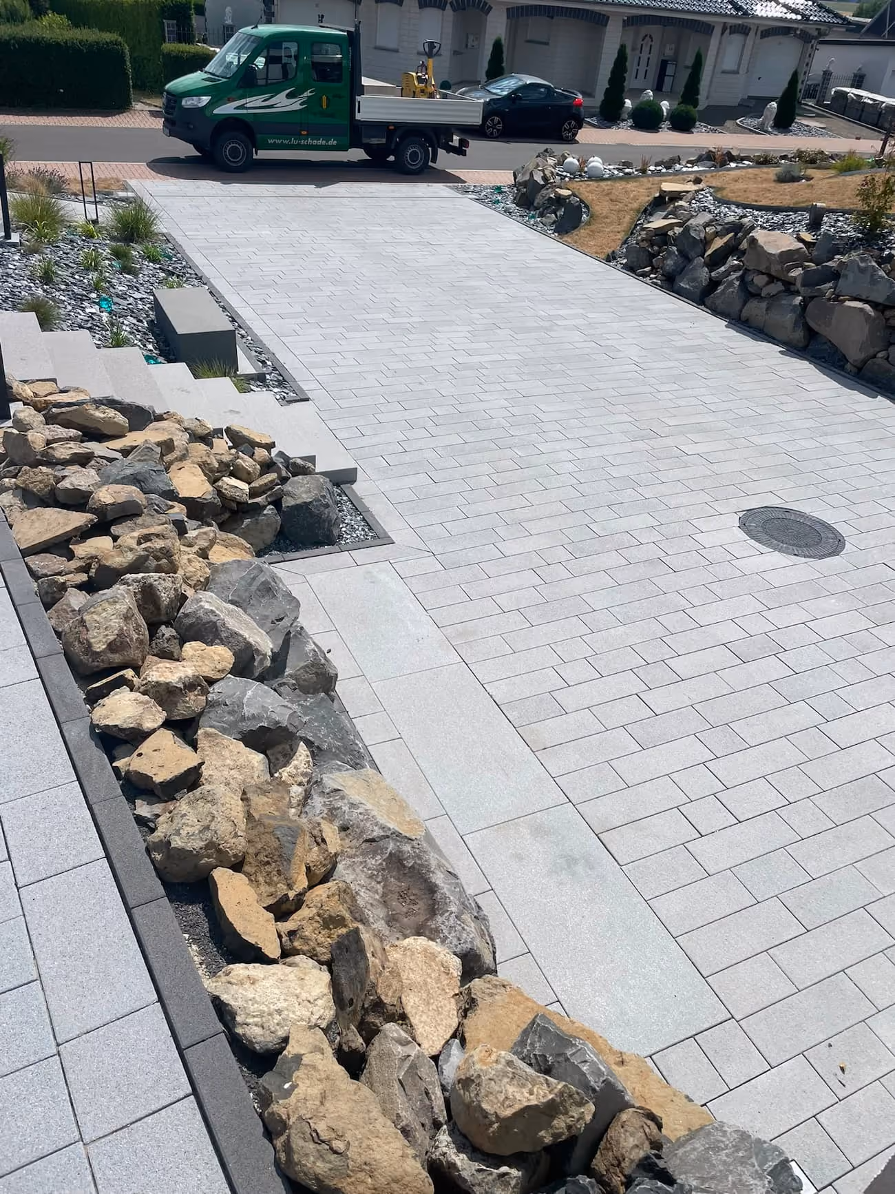 Hellgraue gepflasterte Einfahrt in Homberg (Efze)  mit Felsen und einem grünen Gebr. Schade Firmenkipper am Straßenrand. Garten und Landschaftsbau Referenz von Gebr. Schade 