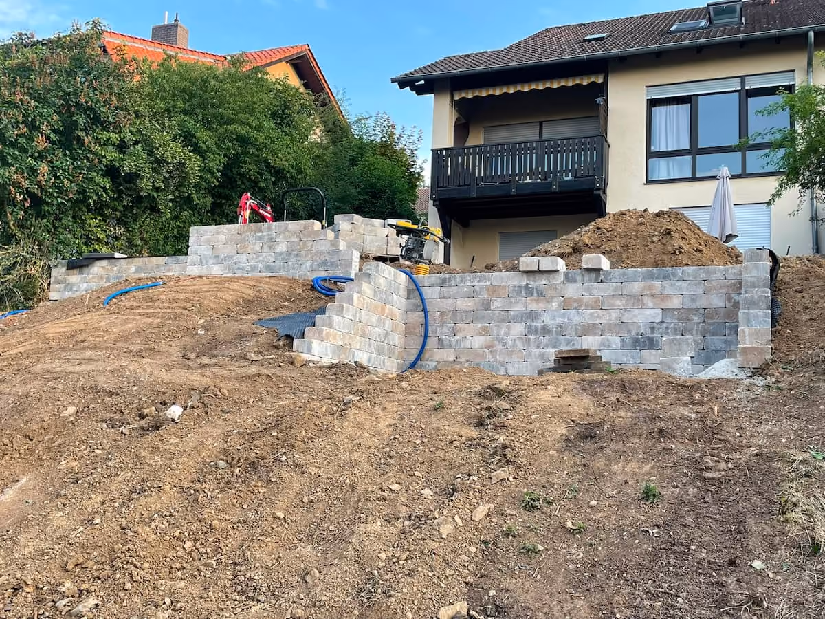Hanggrundstück in Bad Hersfeld mitten im GaLa Bau befindlicher Trockenmauer aus grauen Steinen vor einem Wohnhaus.