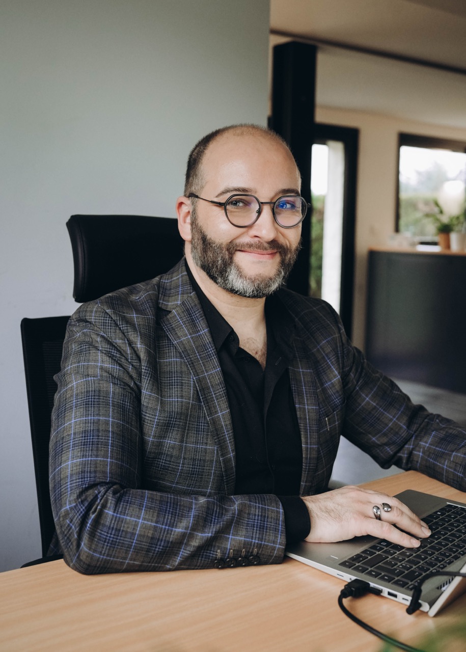 Homme barbe avec lunettes souriant en veste à carreaux travaille sur ordinateur portable à un bureau.