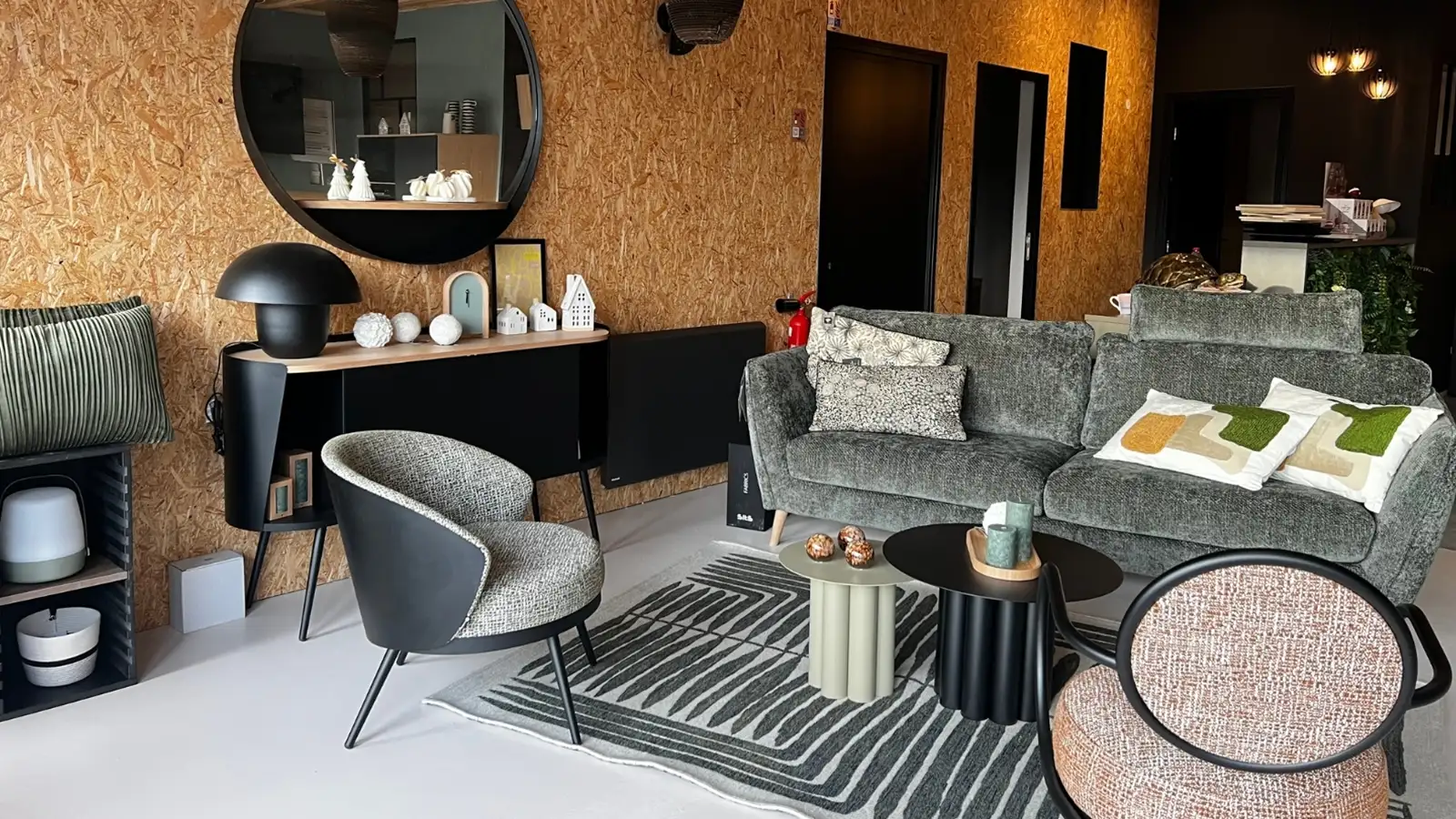 Intérieur du magasin So!cozi avec un salon moderne avec canapé vert, fauteuils à motifs, tapis texturé, miroir rond et mur en bois.