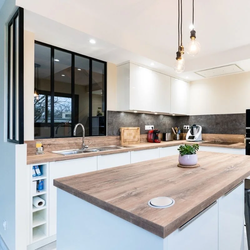 Cuisine blanche avec îlot central en bois, plan de travail effet chêne et verrière intérieure noire, réalisation So!Cozi