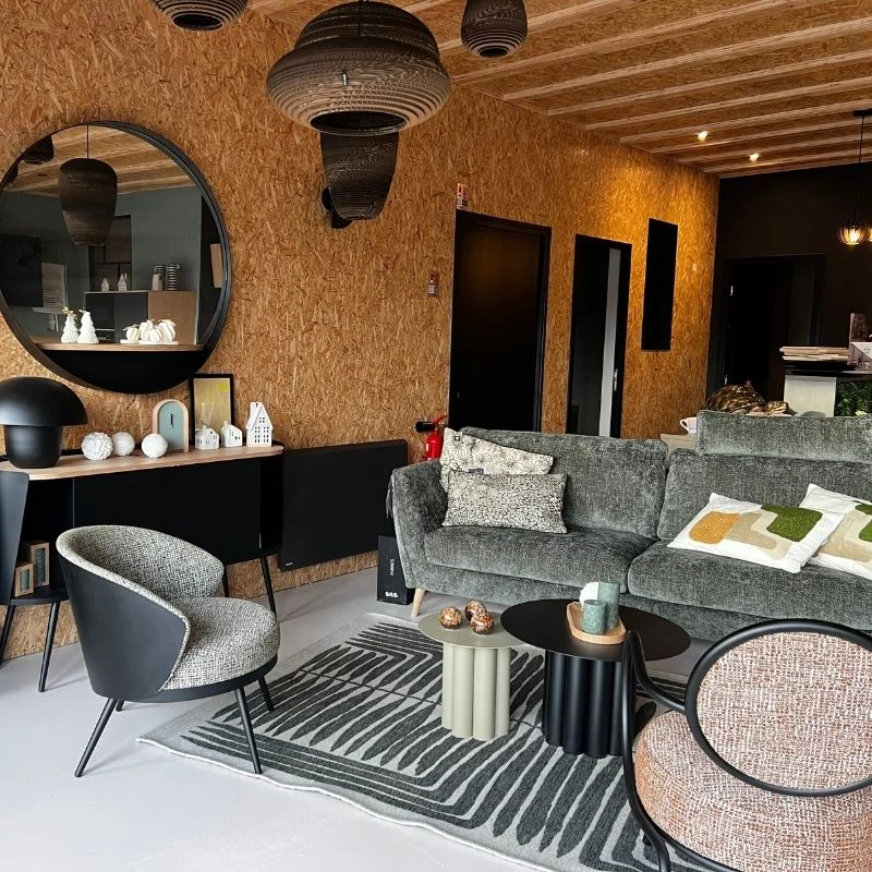 Intérieur du magasin So!cozi avec un salon moderne avec canapé vert, fauteuils à motifs, tapis texturé, miroir rond et mur en bois.