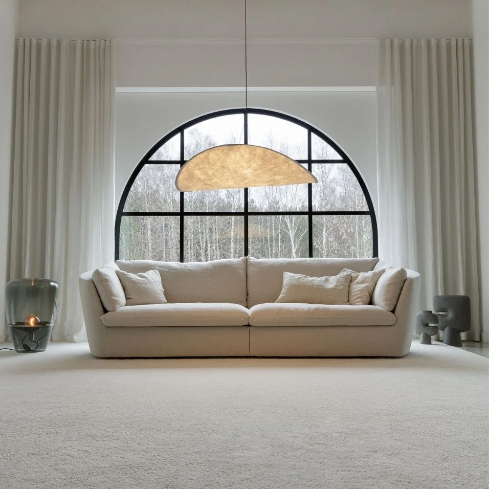 Canapé beige moderne Bonnie de Sits, placé sous une grande fenêtre, avec une suspension lumineuse. Disponible chez So!Cozi