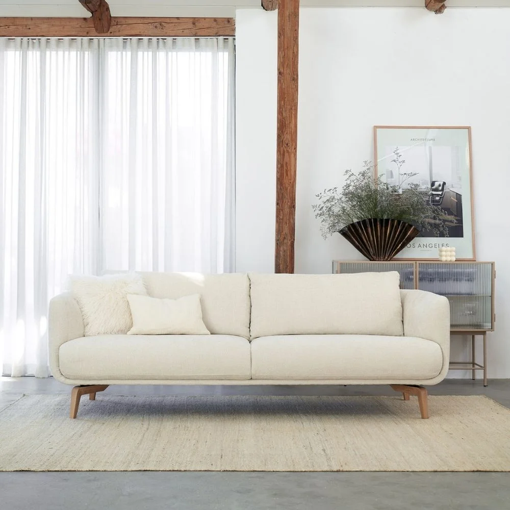 Canapé beige moderne Moa de Sits, posé sur un tapis devant des rideaux blancs et un buffet en bois. Disponible chez So!Cozi