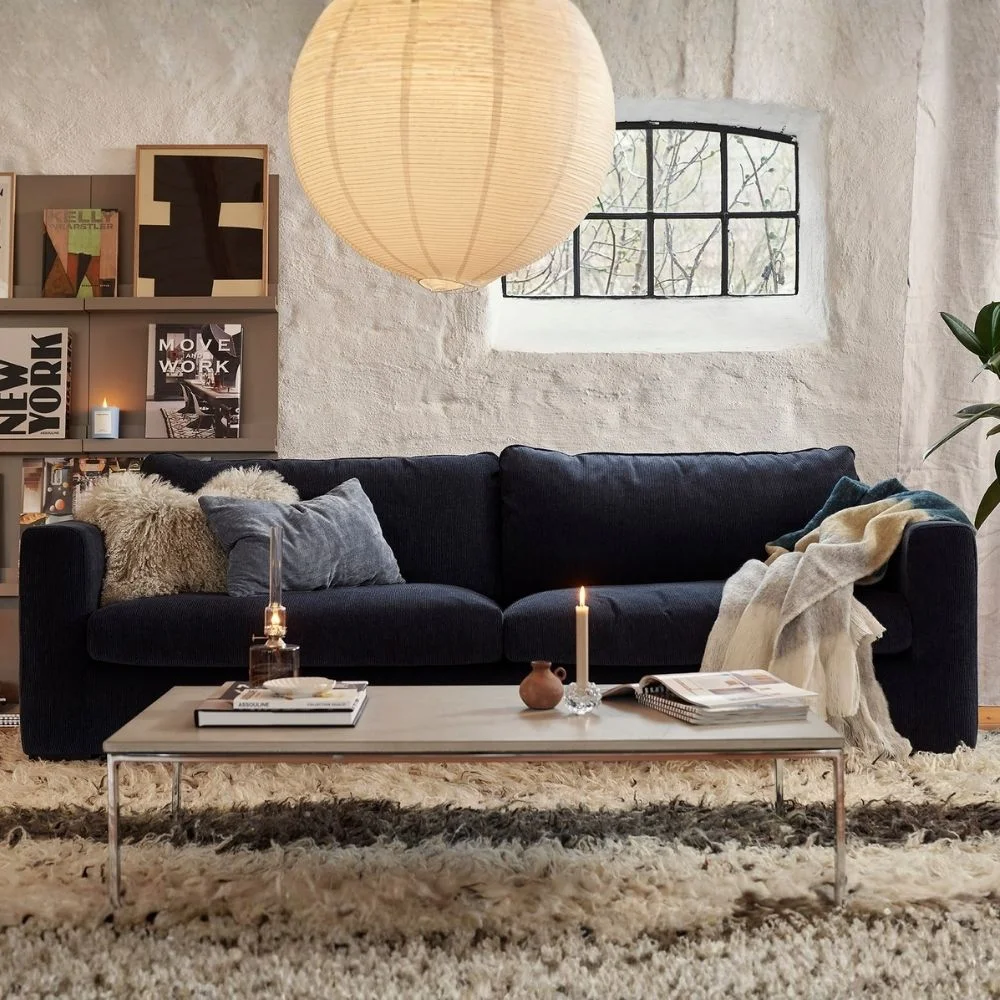 Canapé bleu foncé Sophia de Sits avec des coussins texturés et un plaid, dans une pièce chaleureuse avec une lanterne en papier et d'une œuvre d'art. Disponible chez So!Cozi